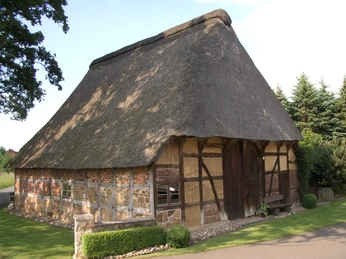 5.05_Handwerksdorf Frotheim__Brammeyers Scheune_LandArt-Station 5.05.JPG Fachwerkgebäude mit Reetdach in ländlicher Umgebung, umgeben von Bäumen und gepflegtem Garten.