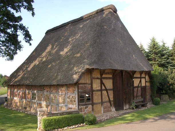 5.05_Handwerksdorf Frotheim__Brammeyers Scheune_LandArt-Station 5.05.JPG Fachwerkgebäude mit Reetdach in ländlicher Umgebung, umgeben von Bäumen und gepflegtem Garten.