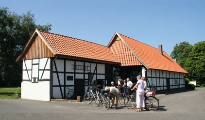 Alte Schule Wietersheim_Radfahrergruppe am Fachwerkhaus.jpg Eine Gruppe von Radfahrern pausiert vor einem traditionellen Fachwerkhaus unter klarem Himmel.