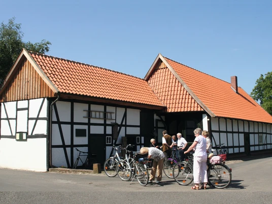 Alte Schule Wietersheim_Radfahrergruppe am Fachwerkhaus.jpg Eine Gruppe von Radfahrern pausiert vor einem traditionellen Fachwerkhaus unter klarem Himmel.