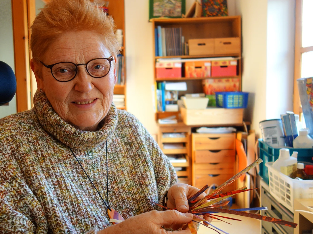 4.28_Kuenstlerin_Bettina Bollmann-Koch QUERformat_Bettina Bollmann-Koch.JPG Künstlerin Bettina Bollmann-Koch im Atelier, lächelnd mit selbst gestalteten Papierstreifen.