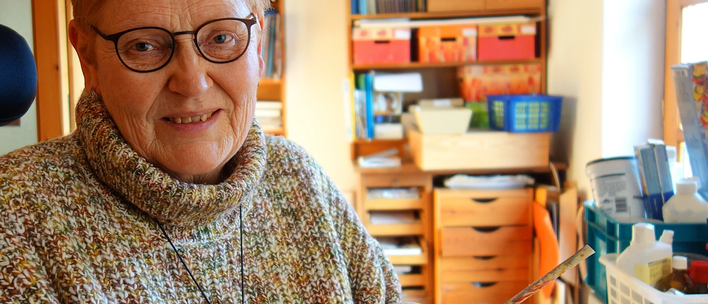 4.28_Kuenstlerin_Bettina Bollmann-Koch QUERformat_Bettina Bollmann-Koch.JPG Künstlerin Bettina Bollmann-Koch im Atelier, lächelnd mit selbst gestalteten Papierstreifen.