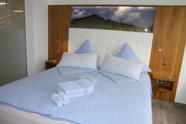 Helles Doppelbett in modernem Zimmer mit zwei blauen Kissen, Handtücher gefaltet am Fußende.
