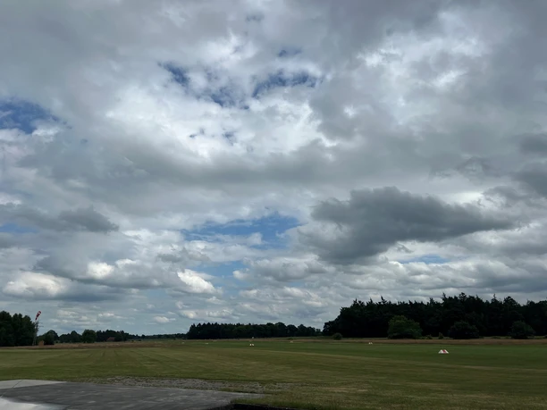 Bild1.jpg Das Bild zeigt eine große, wolkenverhangene Wiese mit bewölktem Himmel. Der Horizont ist von Wäldern gesäumt. Eine ruhige Landschaft bei wechselhaftem Wetter.