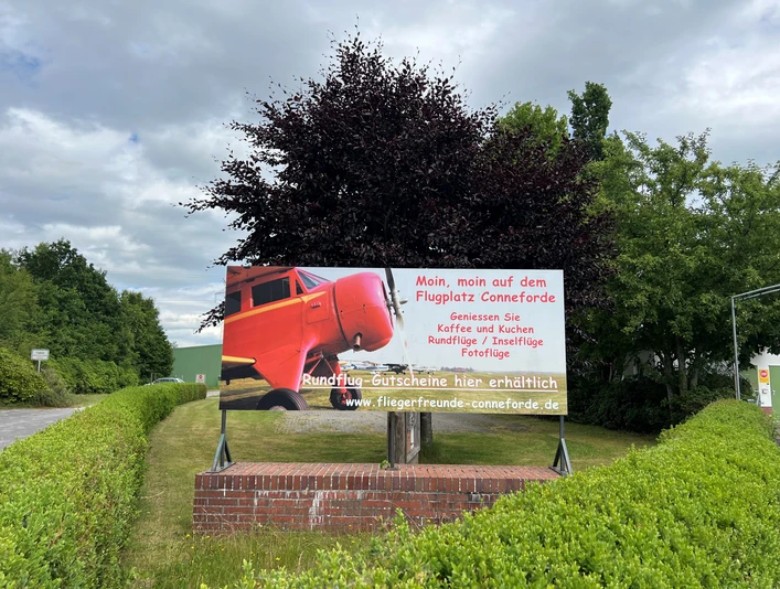 Bild3.jpg Auf dem Bild ist ein Schild am Flugplatz Conneforde zu sehen. Es zeigt ein rotes Flugzeug und wirbt für Rundflüge und Gutscheine der "Fliegerfreunde Conneforde". Umgeben von grünen Hecken steht das Schild vor einem Baum.