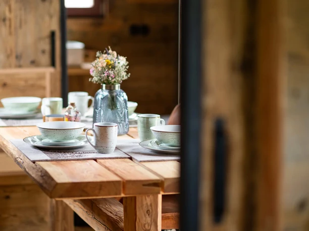 Ein rustikaler Holztisch in einem Tiny House der Harz-Urlaubs-Alm, Buntenbock, ist mit stilvollem Geschirr gedeckt, in der Mitte des Tisches steht ein Blumenarrangement in einer Vase.