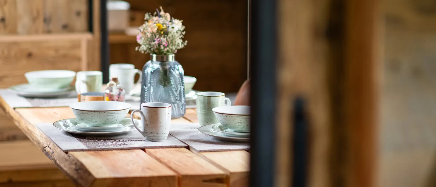 Harz-Urlaubs-Alm in Buntenbock - gedeckter Tisch im Tiny House Ein rustikaler Holztisch in einem Tiny House der Harz-Urlaubs-Alm, Buntenbock, ist mit stilvollem Geschirr gedeckt, in der Mitte des Tisches steht ein Blumenarrangement in einer Vase.