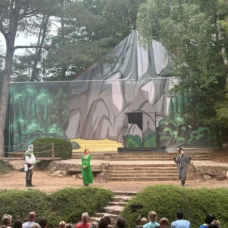 233a758a-ba33-437b-b422-f5d0585a9afc.jpg Freilichtbühne mit Schauspielenden in Kostümen vor einer Kulisse aus Felsen und Bäumen