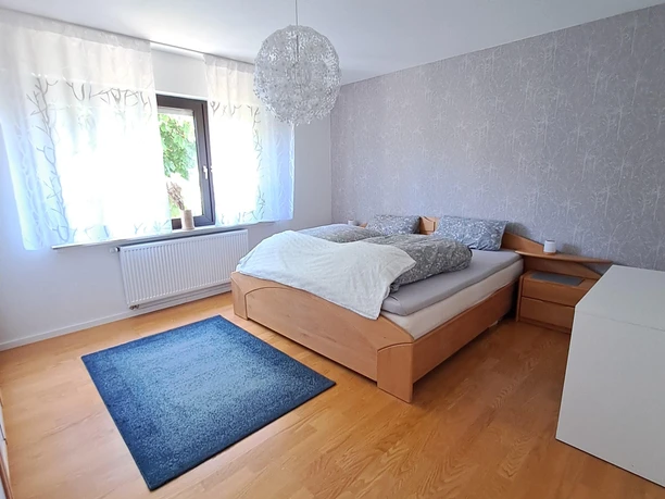 Ein helles Schlafzimmer mit Holzbett, blauen Teppich und einem Fenster mit Blick auf grüne Bäume.