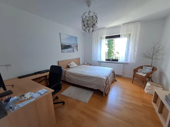 Ein helles Schlafzimmer mit Doppelbett, Schreibtisch, Stuhl, Fenster mit Vorhang und einem Sessel.