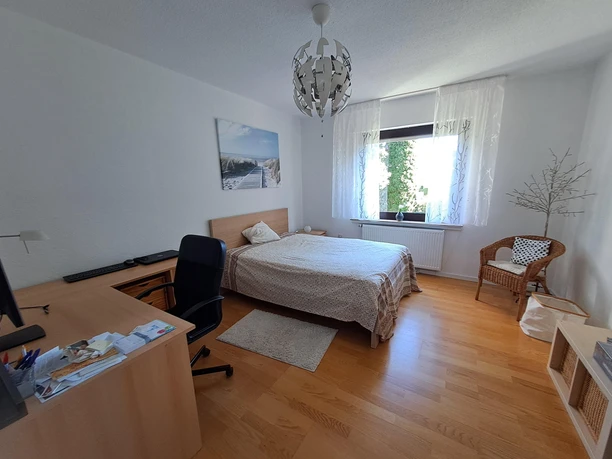 Ein helles Schlafzimmer mit Doppelbett, Schreibtisch, Stuhl, Fenster mit Vorhang und einem Sessel.