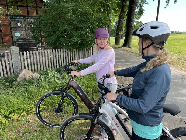 Two women wearing cycle helmets take a break on e-bikes beside a dirt track, next to trees.