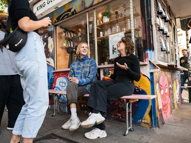 King Georg Büdchen Das Bild zeigt eine lebhafte Szene vor einem farbenfroh gestalteten Kiosk. Eine kleine Gruppe junger Menschen ist in entspannter Unterhaltung vertieft. Im Vordergrund sitzen zwei Personen auf einer Bank und genießen die lebendige Atmosphäre des Großstadtflairs.