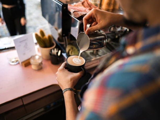 King Georg Büdchen Barista gießt kunstvoll Milch in einen Kaffee, der von einer Person in einem bunt karierten Hemd gehalten wird.