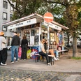 King Georg Büdchen Kleines Kiosk im urbanen Umfeld, umgeben von Bäumen und Menschen in entspannter Atmosphäre.Small kiosk in an urban setting, surrounded by trees and people in a relaxed atmosphere.
