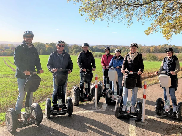 Tour Herford 1 (2)_.jpg Gruppe von Menschen auf Segways fährt bei sonnigem Wetter entlang eines Feldwegs, im Hintergrund Wälder.