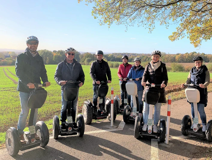Tour Herford 1 (2)_.jpg Gruppe von Menschen auf Segways fährt bei sonnigem Wetter entlang eines Feldwegs, im Hintergrund Wälder.