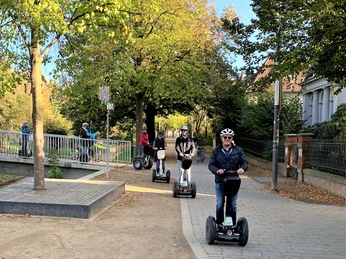 Segway Tour Herford_SEGBI (7).jpg Eine Gruppe von Menschen fährt mit Segways auf einem gepflasterten Weg durch eine grüne Parklandschaft.