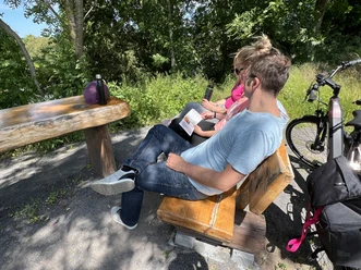 A couple is sitting on a wooden bench in a shady park, bicycles parked next to them.