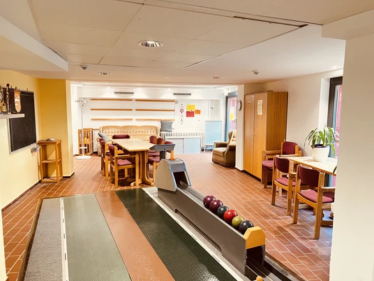 Kegelbahn (1).JPG Kegelbahn im Innenbereich mit Tischen und Stühlen im Hintergrund, helle Beleuchtung, Pflanze am Rand.