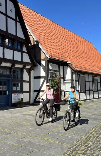 Zwei Radfahrer in sportlicher Kleidung vor historischen Fachwerkhäusern unter strahlend blauem Himmel.