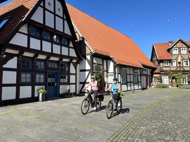 Two cyclists in sporty clothing in front of historic half-timbered houses under a bright blue sky.