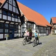 Radfahren in der Mittelweser-Region Zwei Radfahrer in sportlicher Kleidung vor historischen Fachwerkhäusern unter strahlend blauem Himmel.