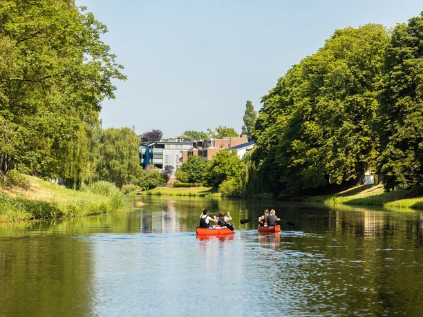 Zwei rote Kanus mit Menschen auf einem ruhigen Fluss, umgeben von üppigen grünen Bäumen.