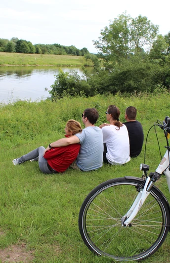 Vier Menschen genießen die Aussicht auf einen Fluss, während zwei Fahrräder auf einer Wiese stehen.
