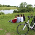 Radfahren in der Mittelweser-Region Vier Menschen genießen die Aussicht auf einen Fluss, während zwei Fahrräder auf einer Wiese stehen.