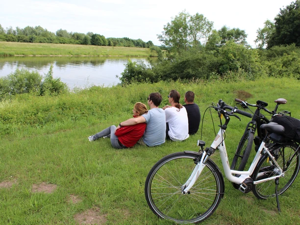 Radfahren in der Mittelweser-Region Vier Menschen genießen die Aussicht auf einen Fluss, während zwei Fahrräder auf einer Wiese stehen.