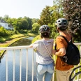 herford-promenade-teutoburger-wald-tourismus-h-tornow-018-jpg.jpg Zwei Radfahrer mit Helmen stehen auf einer Brücke und blicken in einen sonnigen, grünen Park.