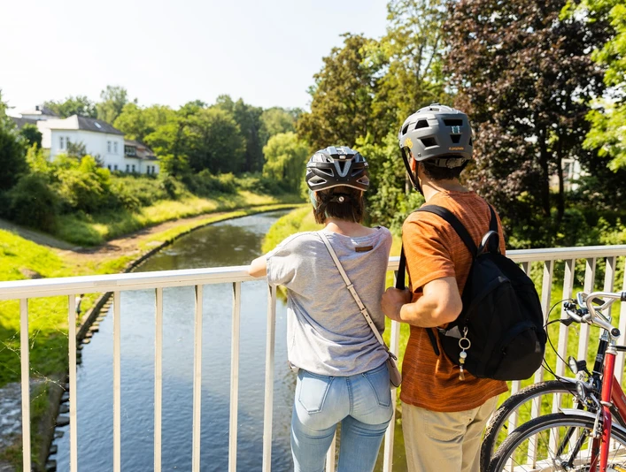herford-promenade-teutoburger-wald-tourismus-h-tornow-018-jpg.jpg Zwei Radfahrer mit Helmen stehen auf einer Brücke und blicken in einen sonnigen, grünen Park.