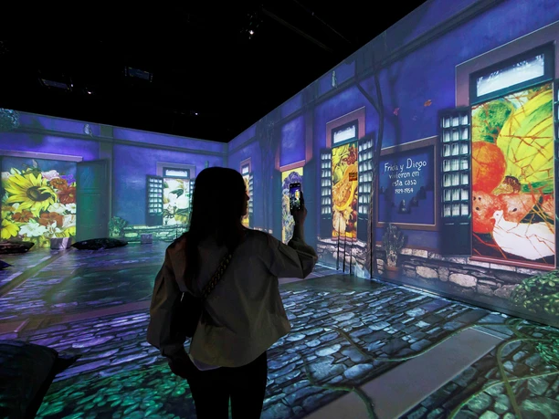 VIVA FRIDA KAHLO The picture shows a person in a dark room, surrounded by luminous projections with motifs by the artist Frida Kahlo on the walls and floor, creating an immersive art experience.