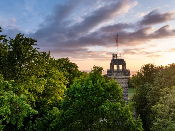 Herford-Bismarckturm-2025-067-©Teutoburger Wald - WfG Herford - D. Ketz.jpg Bismarckturm Herford im Abendlicht, umgeben von grünem Laub und einem dramatischen Wolkenhimmel.