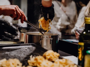 MarkthalleHerford-81.jpg Koch in schwarzem Handschuh gibt frische Pasta mit einer Zange in einen Topf mit dampfendem Wasser.