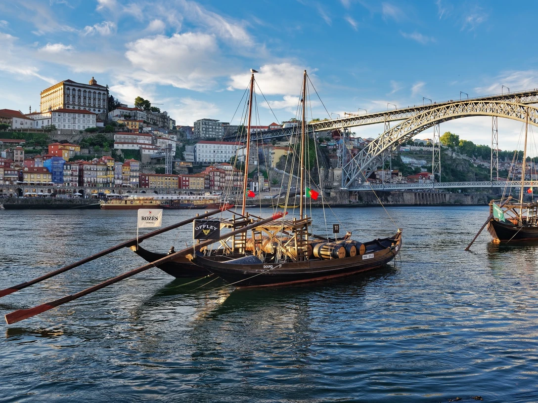 Portugal_Kal. SHS.jpg Das Bild zeigt traditionelle Boote im Fluss Douro vor der beeindruckenden Kulisse Portos. Im Hintergrund erhebt sich die ikonische Dom-Luís-I-Brücke vor einer üppigen Flusslandschaft.