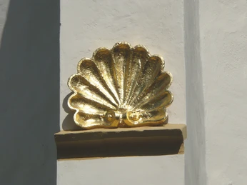 Pilgersymbol an St Jakobi Herford - ProHerford GmbH.jpg Goldene Pilgermuschel in Herford, Symbol des Jakobsweges, kontrastiert vor weißer Wand.