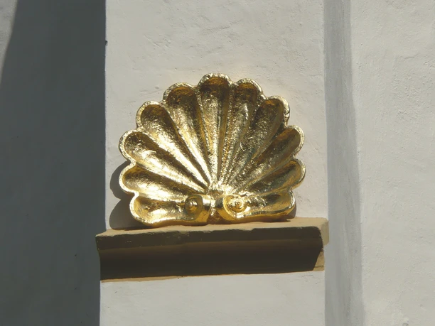 Pilgersymbol an St Jakobi Herford - ProHerford GmbH.jpg Goldene Pilgermuschel in Herford, Symbol des Jakobsweges, kontrastiert vor weißer Wand.