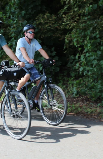 Zwei Männer auf Fahrrädern fahren mit Helmen auf einem asphaltierten Weg, umgeben von Bäumen.