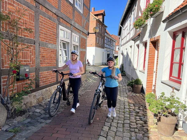 Radfahren in der Mittelweser-Region Zwei Frauen schieben Fahrräder durch eine enge, gepflasterte Gasse mit Fachwerkhäusern und Sonnenschein.