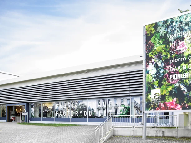 Moderne Gebäudefassade des Ahlers Fashion Outlet Herford mit großen Glasfronten und Plakat.