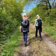 Twee vrouwen met rugzakken lopen over een groen bospad onder een strakblauwe hemel.