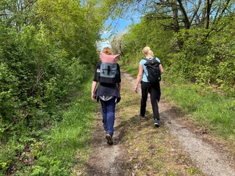 Zwei Frauen mit Rucksäcken wandern auf einem grünen Waldpfad unter klarem, blauen Himmel.