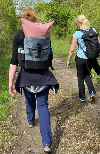 Zwei Frauen wandern auf einem schmalen Waldweg in sommerlichen Outfits und tragen Rucksäcke.