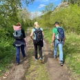 Drei Personen wandern auf einem naturbelassenen Weg, umgeben von frischem Grün und blauem Himmel.
