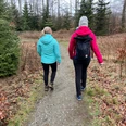 Zwei Menschen in farbigen Jacken und Rucksäcken wandern in einem Waldgebiet auf einem schmalen Pfad.