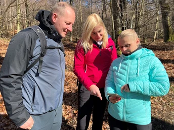 Drei Wanderer in bunter Outdoor-Kleidung studieren ein Smartphone in einem lichten Wald im Herbst.