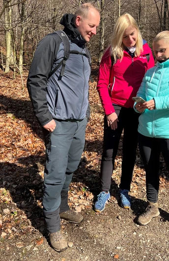 Drei Personen stehen auf einem Waldweg in herbstlichem Laub, betrachten interessiert ein Smartphone.