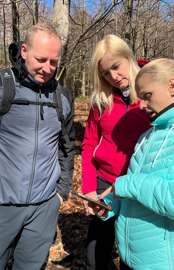 Drei Personen im herbstlichen Wald, fokussiert auf ein gemeinsames Handy; blauer Himmel im Hintergrund.
